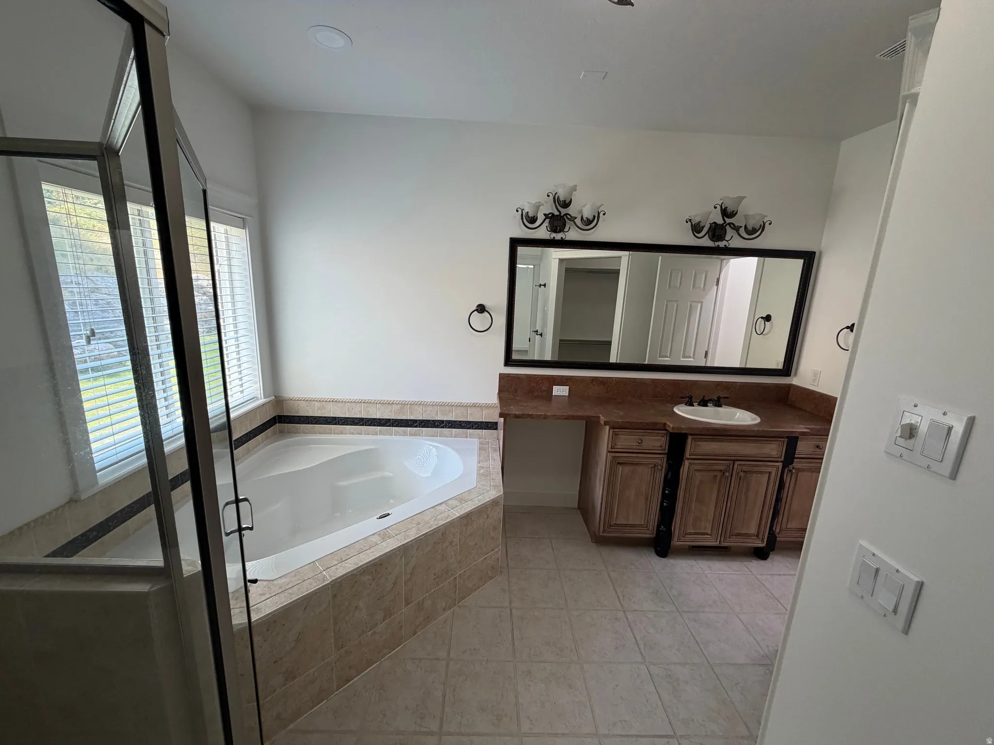 Bathroom featuring a corner soaking tub with tiled surround, a glass-enclosed shower, dual windows with blinds, a vanity with an integrated sink and stone countertop, and an expansive mirror with decorative sconces