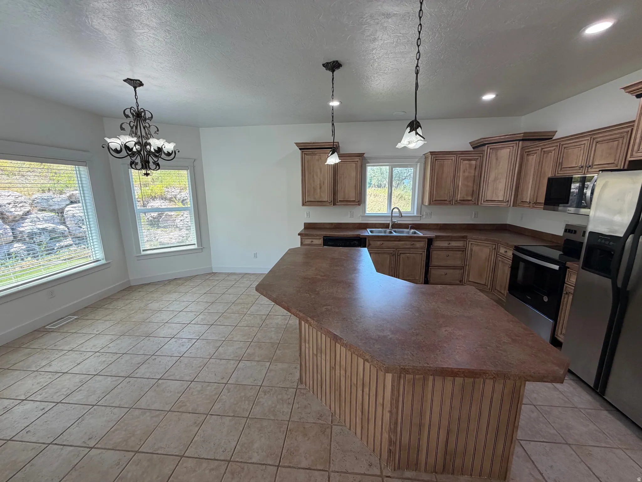 Kitchen and dining area featuring tile flooring, light wood cabinetry, stainless steel appliances, a corner sink with a window, and a chandelier in the dining space