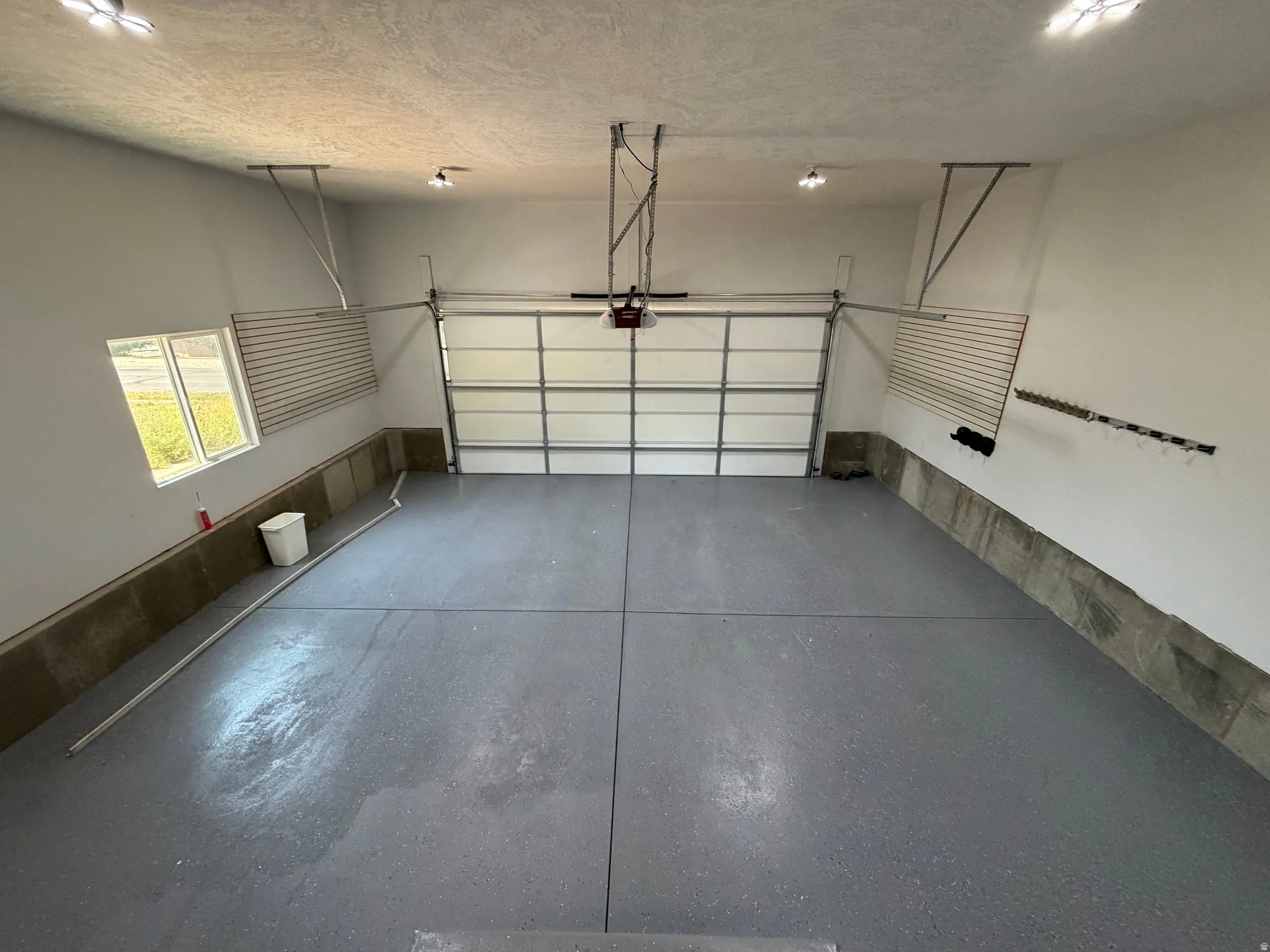 Spacious garage featuring a grey epoxy-coated floor, automatic overhead door, multiple ceiling-mounted light fixtures, and a window providing natural illumination