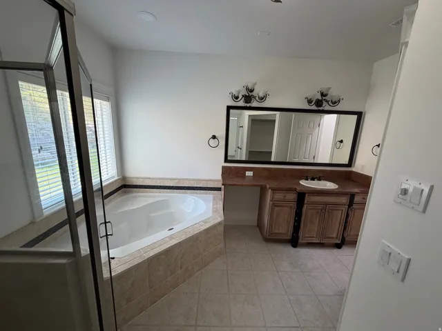 Bathroom featuring a corner soaking tub with tiled surround, a glass-enclosed shower, dual windows with blinds, a vanity with an integrated sink and stone countertop, and an expansive mirror with decorative sconces