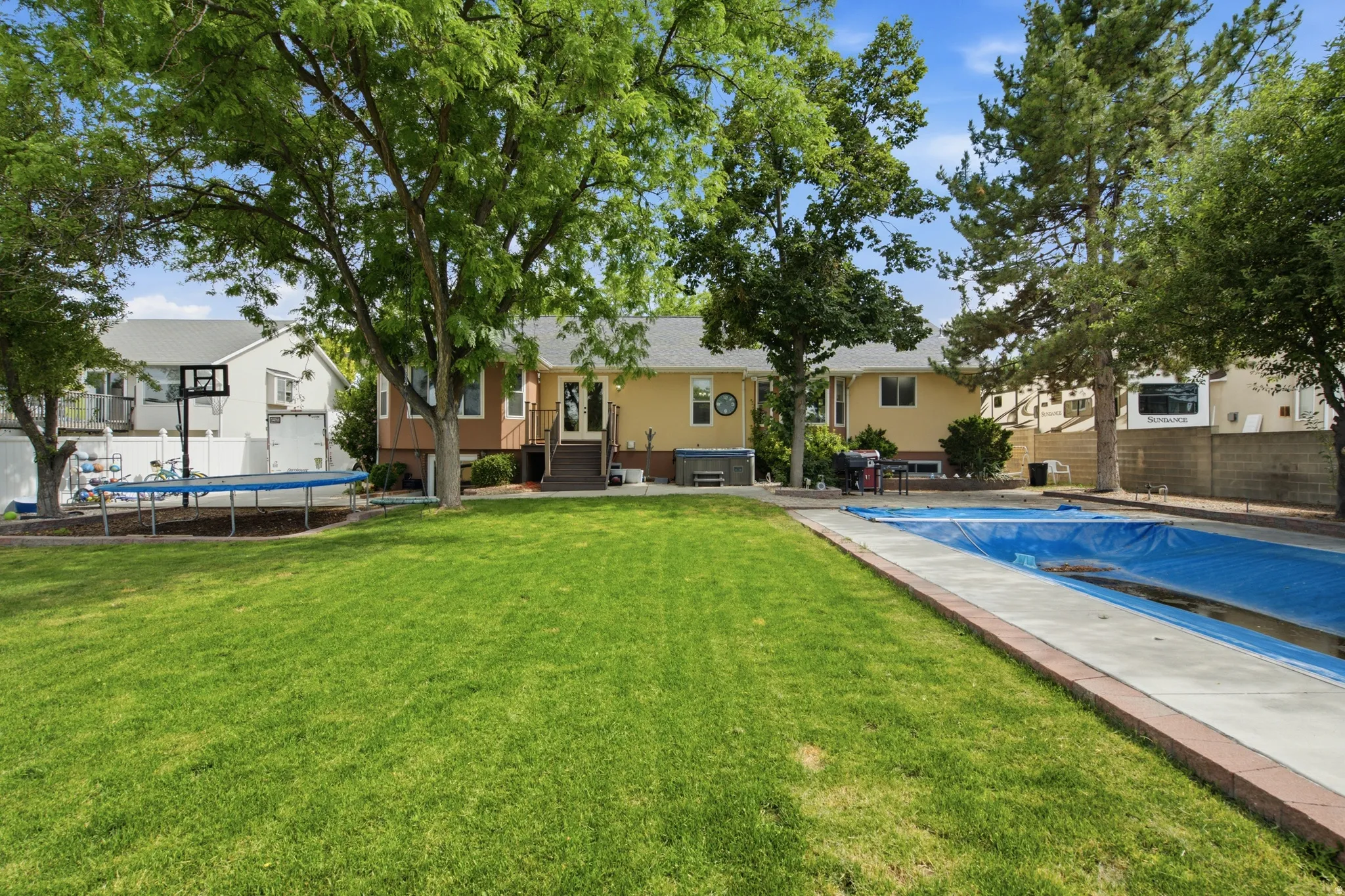 Expansive backyard featuring a covered inground swimming pool, concrete pool deck with brick border, large green lawn, and mature trees providing shade