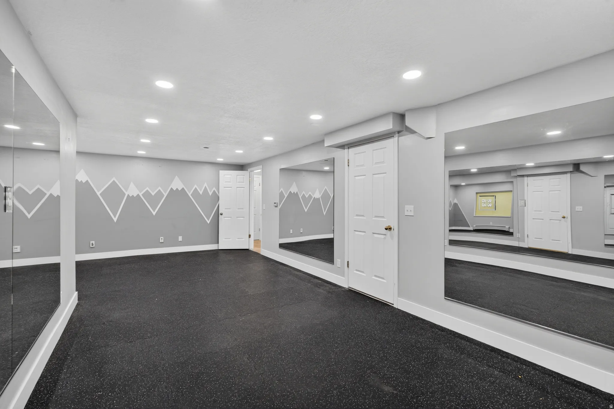 Spacious flexible-use room featuring dark-toned flooring, recessed ceiling lighting, full-height wall mirrors, light gray walls with a mountain motif, and white paneled doors