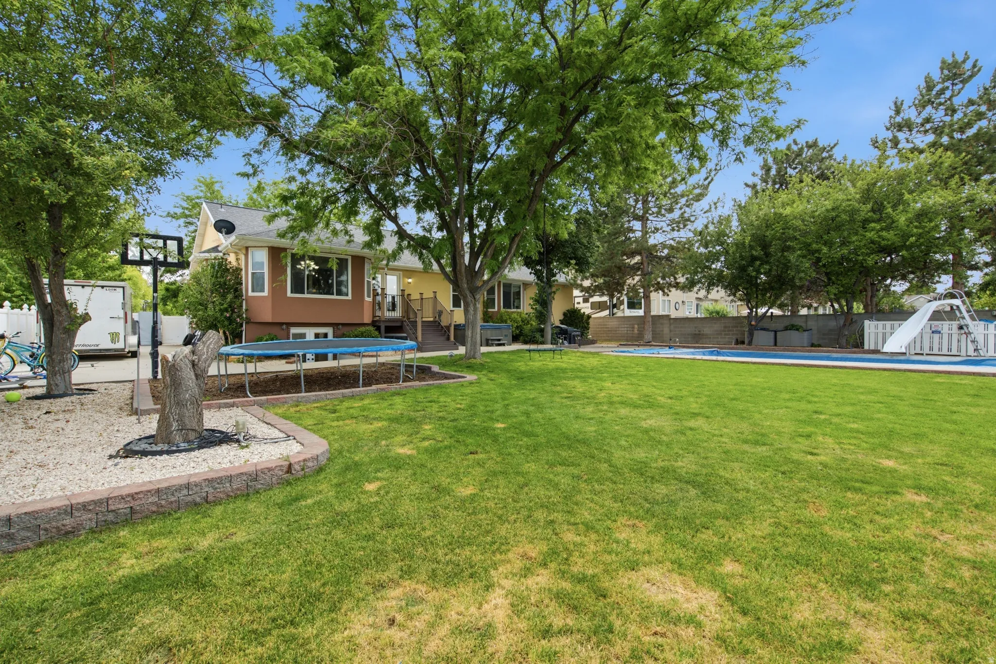 Spacious backyard featuring a large lawn, mature trees, a designated gravel area with a basketball hoop, and a fenced swimming pool