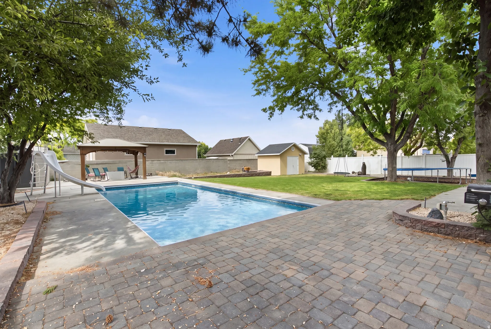 Expansive backyard featuring a rectangular swimming pool with concrete surround, paver patio, a wooden gazebo, and mature trees