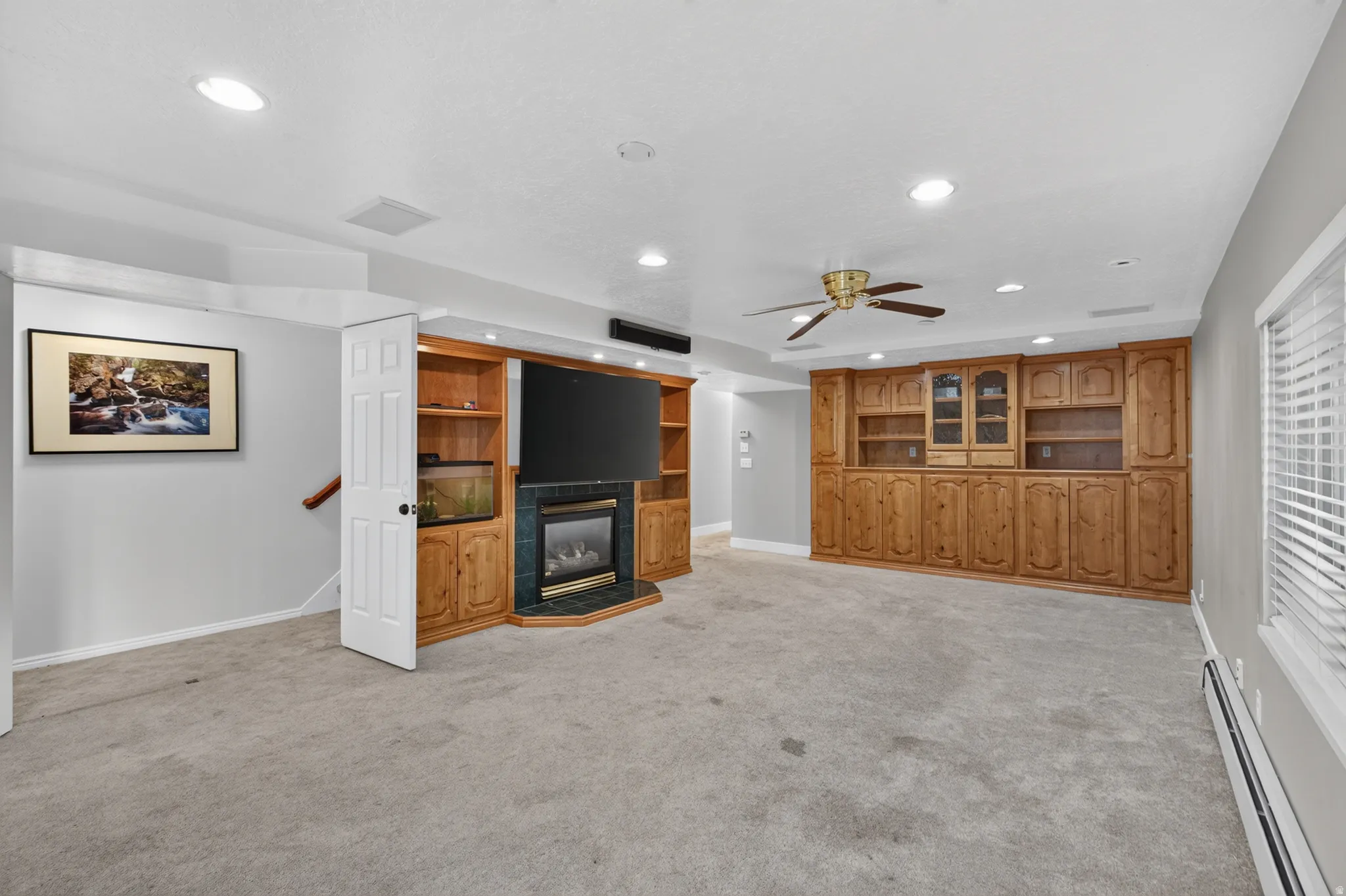 Spacious entertainment room featuring extensive built-in wood cabinetry, a corner-set fireplace with dark stone surround, recessed lighting, and a ceiling fan