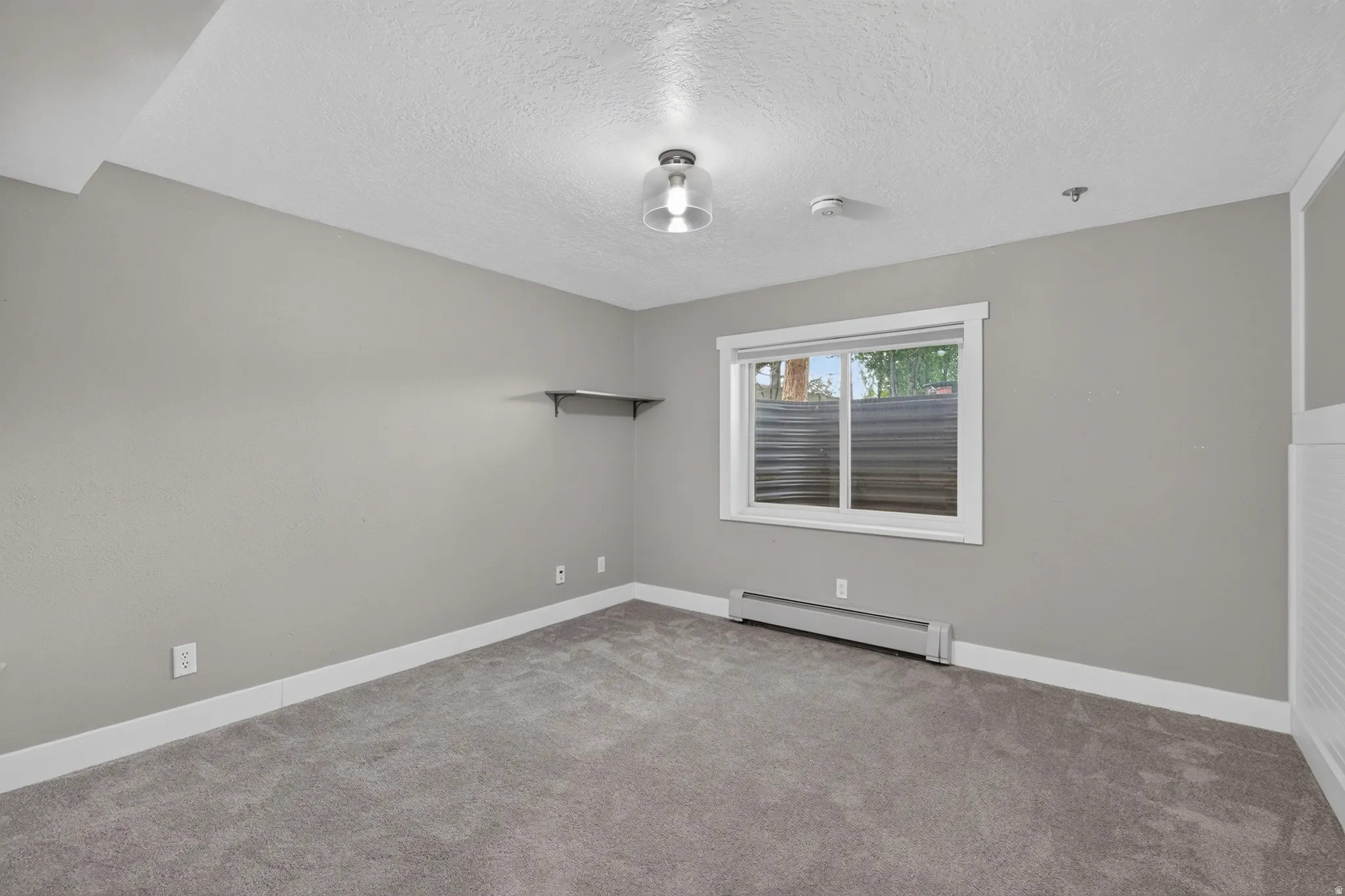 Carpeted room featuring light gray walls, a window with white trim, baseboard heating, and a built-in wall shelf
