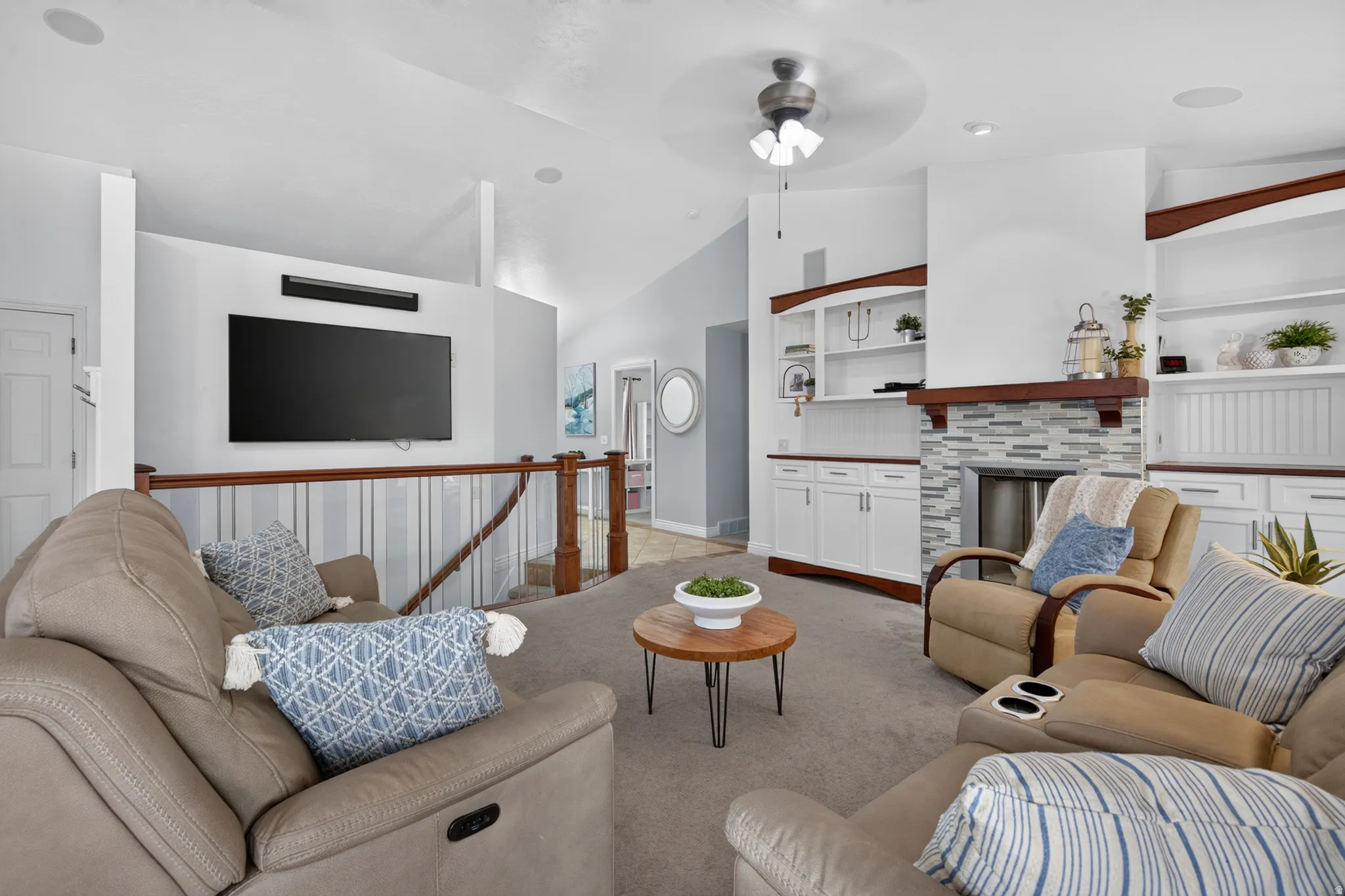 Living area featuring vaulted ceilings, built-in cabinetry with display shelving, a fireplace with a mosaic tile surround, wood-finish railing, and ceiling fan