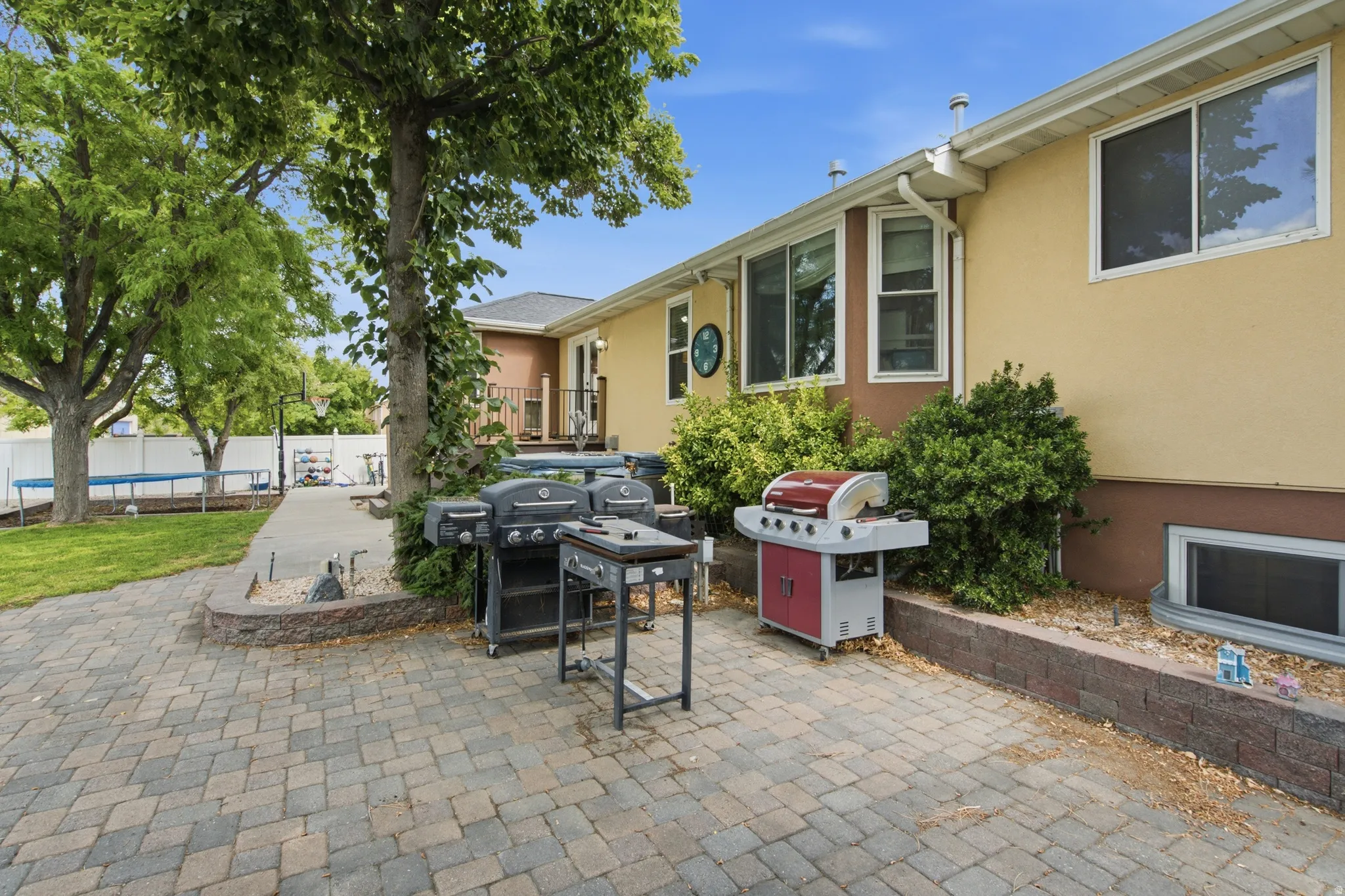Expansive outdoor patio featuring paver stones, a raised brick planter bed, and mature trees providing natural shade