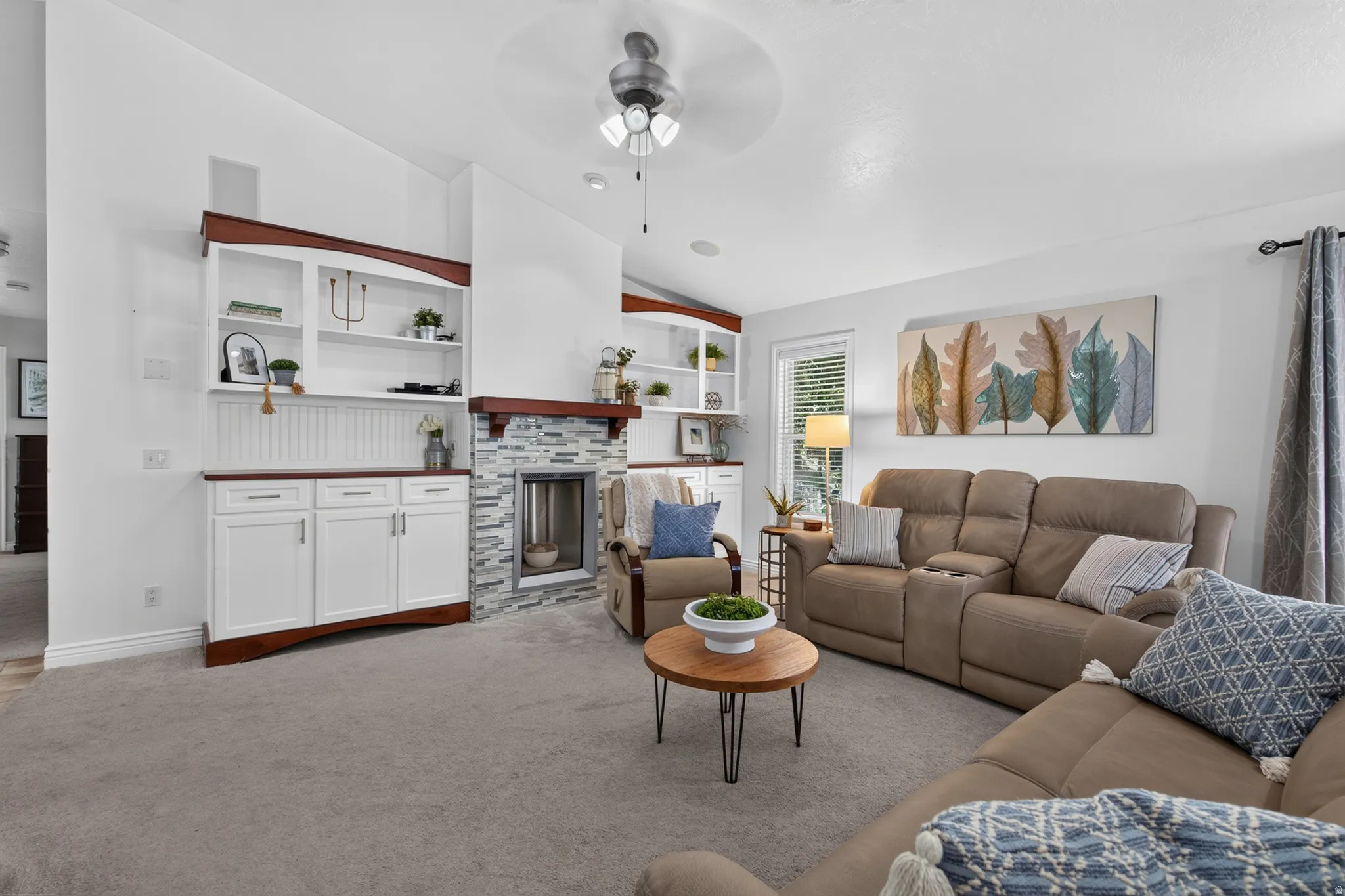 Living area featuring a stone-clad fireplace with a wood mantel, built-in white cabinetry with wood-finish tops, open shelving, and a ceiling fan