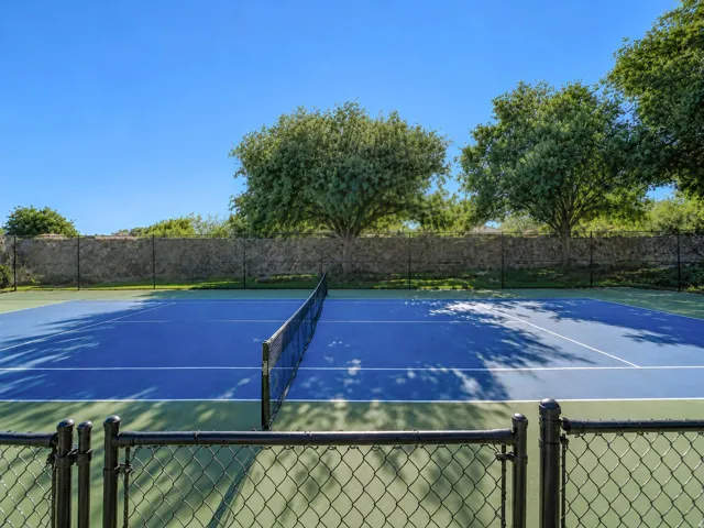 Fenced tennis court featuring a blue playing surface with white lines, surrounded by a stone wall and mature trees