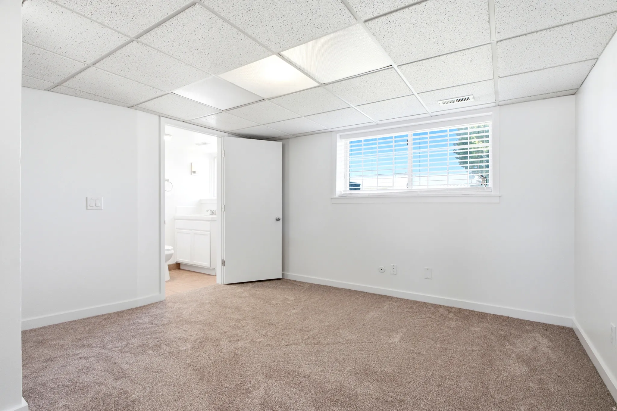 Carpeted room featuring a white interior, drop ceiling with integrated lighting, and a white-framed window with blinds