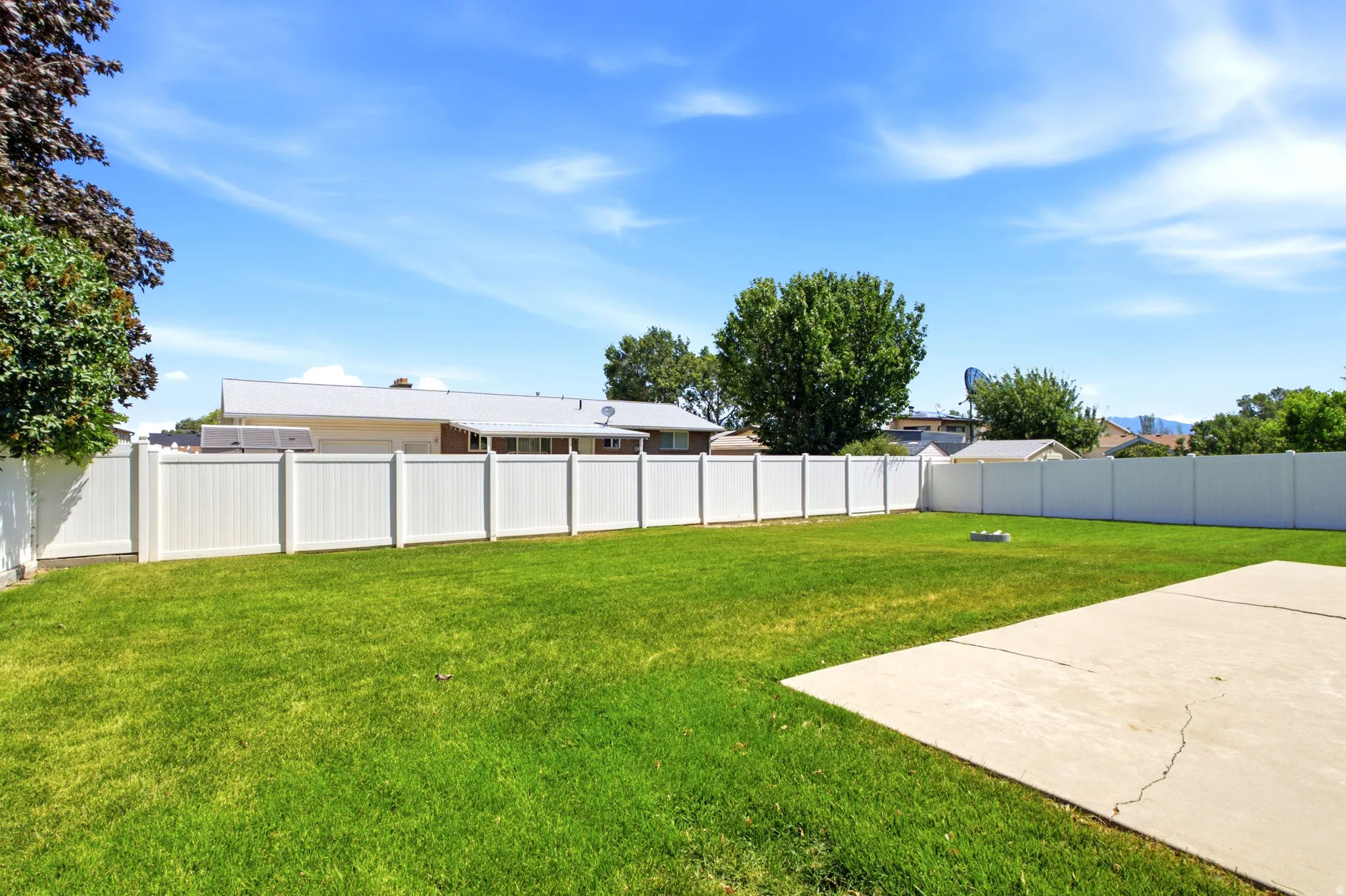 Expansive lawn area featuring a concrete patio, white privacy fencing, and mature trees
