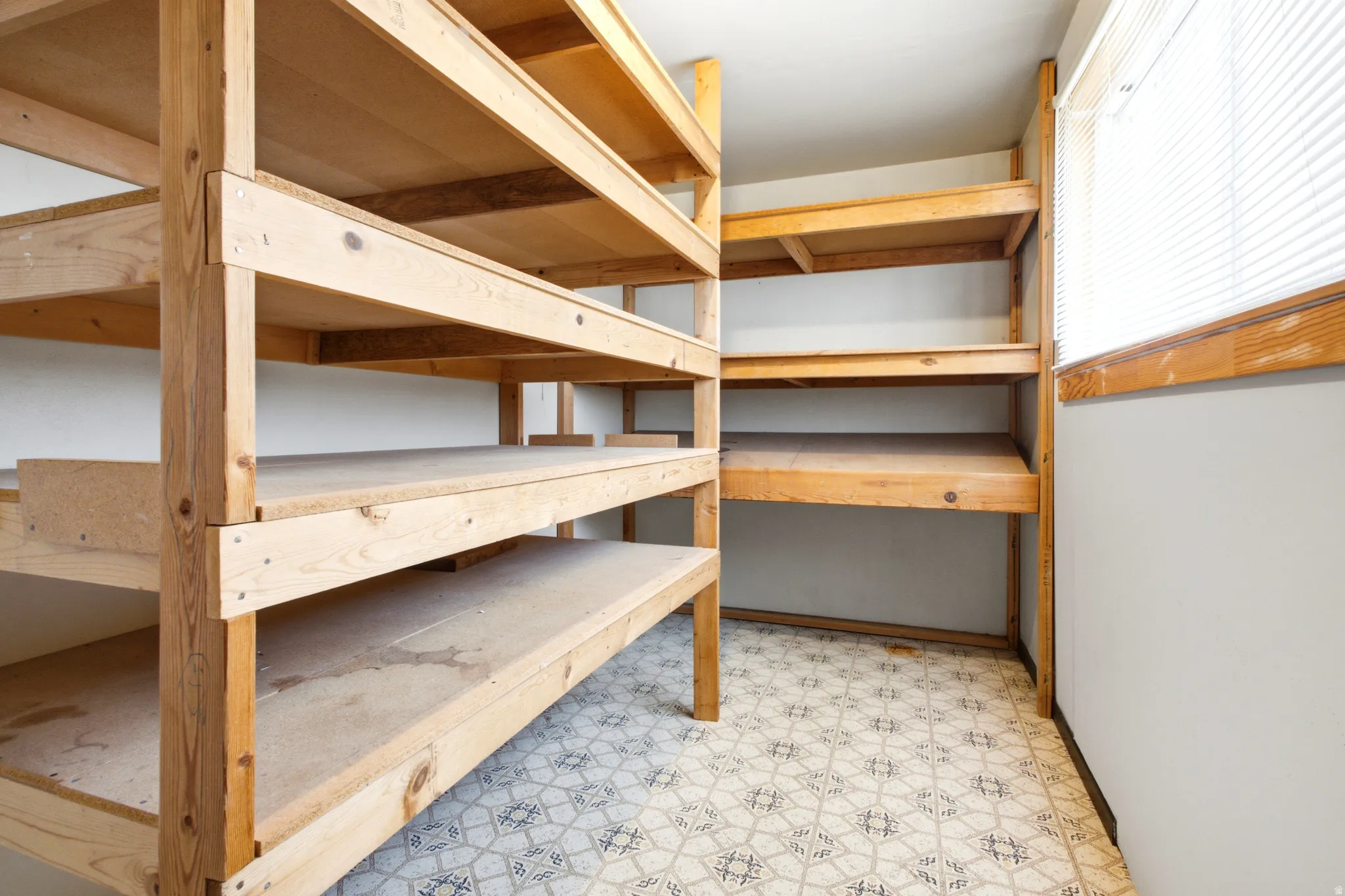 Functional storage room featuring extensive built-in wooden shelving, a patterned floor, and a window with horizontal blinds