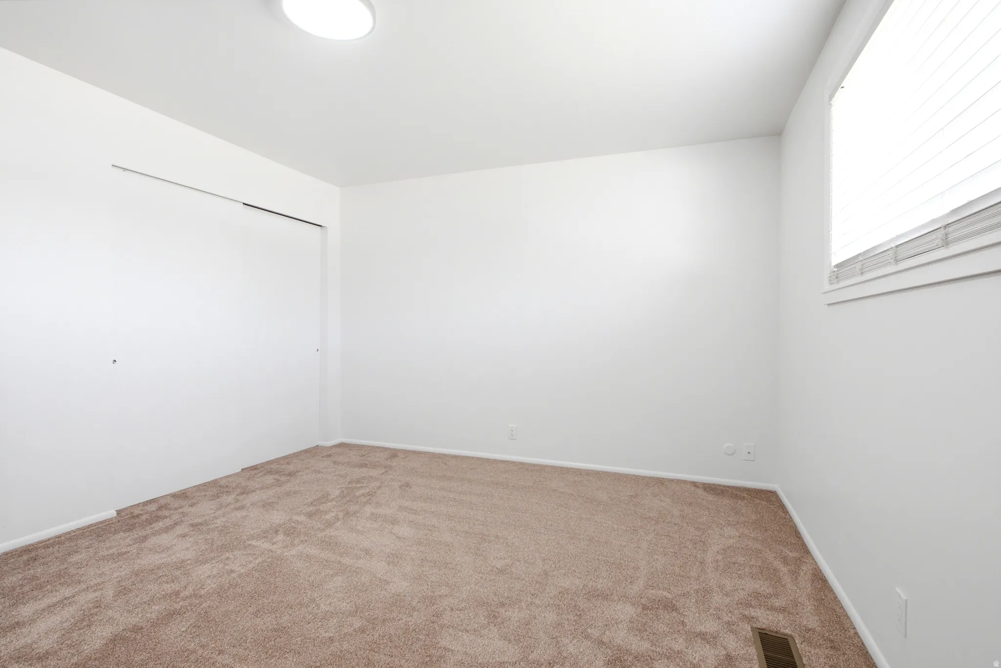 Light-filled room featuring neutral-toned carpeting, white walls, a large window with blinds, a flush-mount ceiling light, and a built-in closet with sliding doors