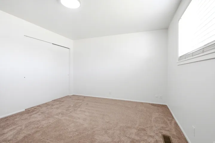 Light-filled room featuring neutral-toned carpeting, white walls, a large window with blinds, a flush-mount ceiling light, and a built-in closet with sliding doors