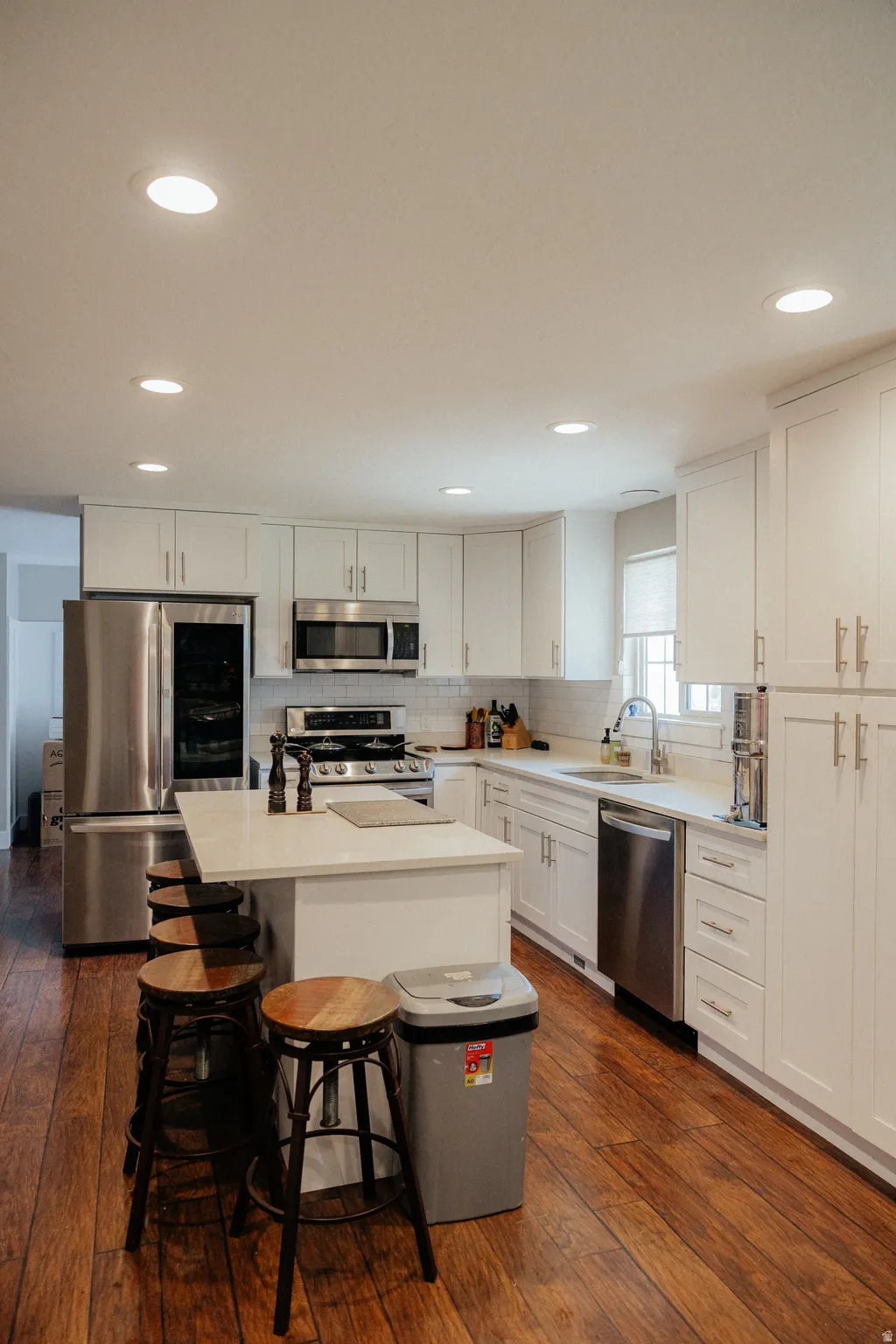 Bright kitchen features abundant white cabinetry, stainless steel appliances, a center island with counter seating, wood-finish flooring, and recessed lighting.