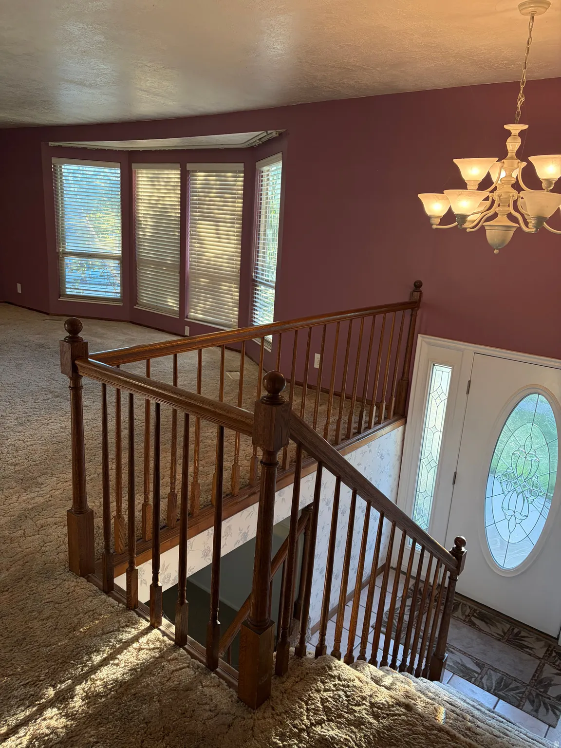 Bay window with blinds, wood newel posts and balusters, chandelier with frosted glass shades, main entry door with oval stained glass, and decorative tile flooring