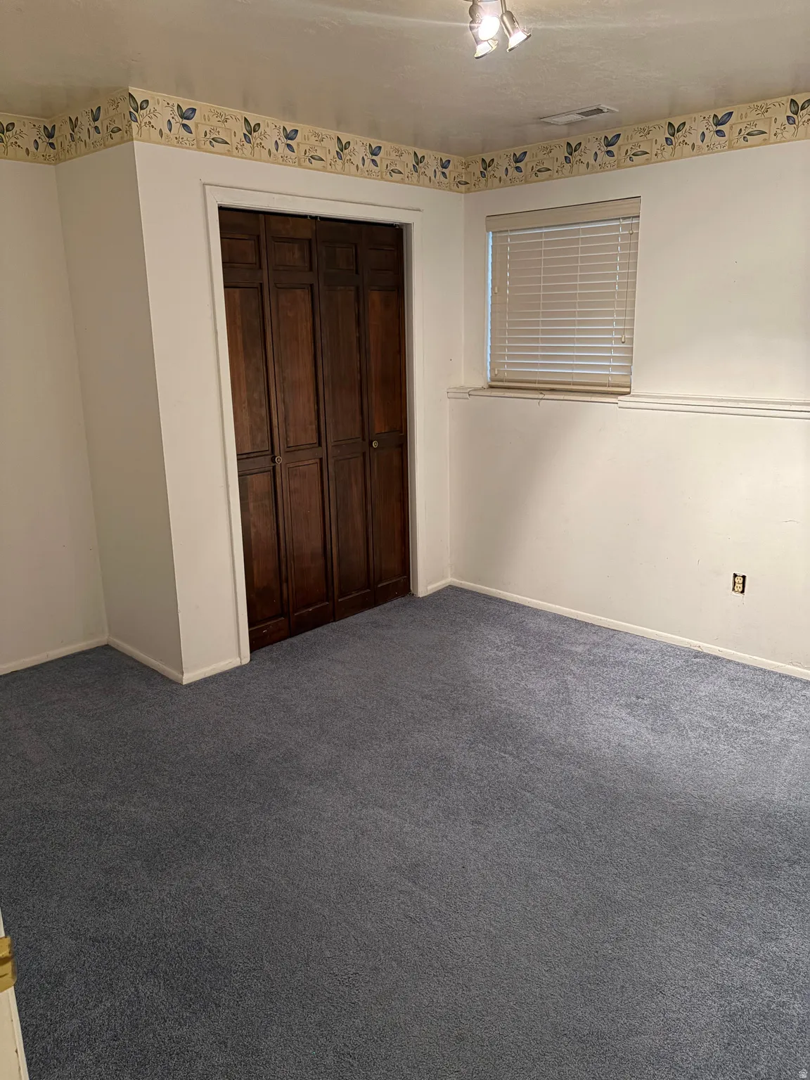 Recessed window with blinds, dark wood-paneled bi-fold closet doors, wall-mounted track lighting, and blue carpeting