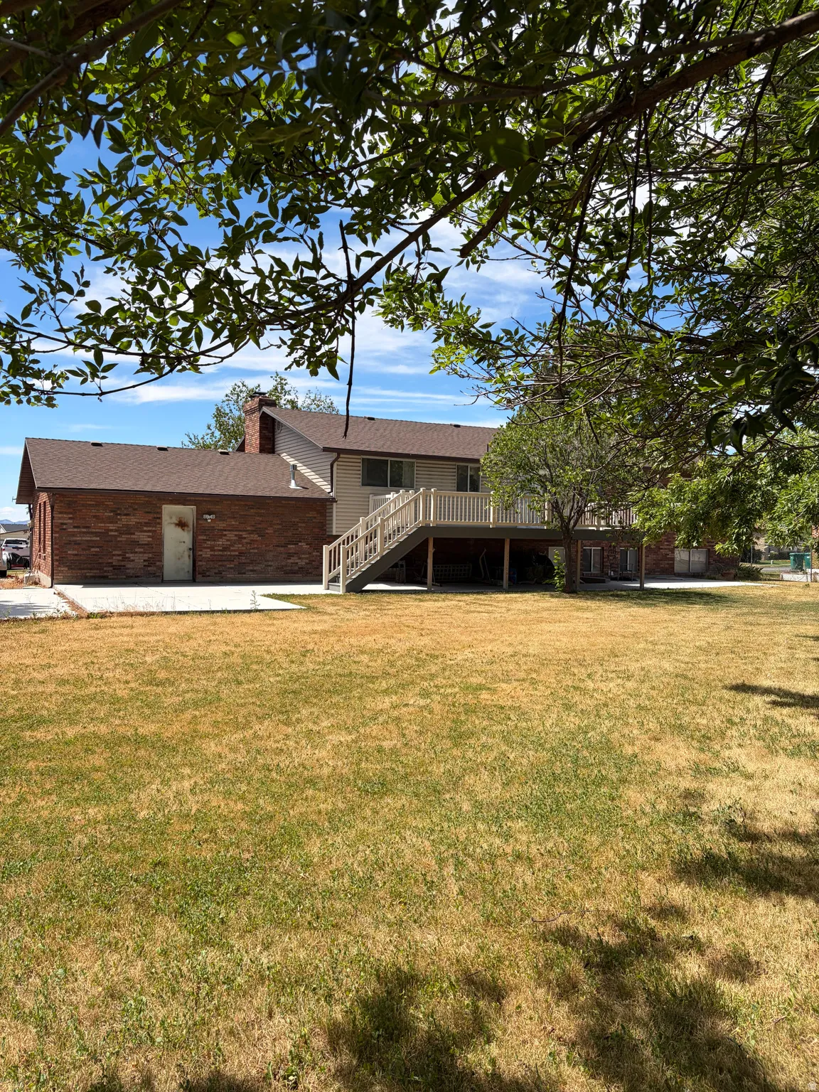 Expansive backyard featuring a large green space, a two-story residence with a brown shingle roof, a brick exterior section, and an elevated deck with a staircase