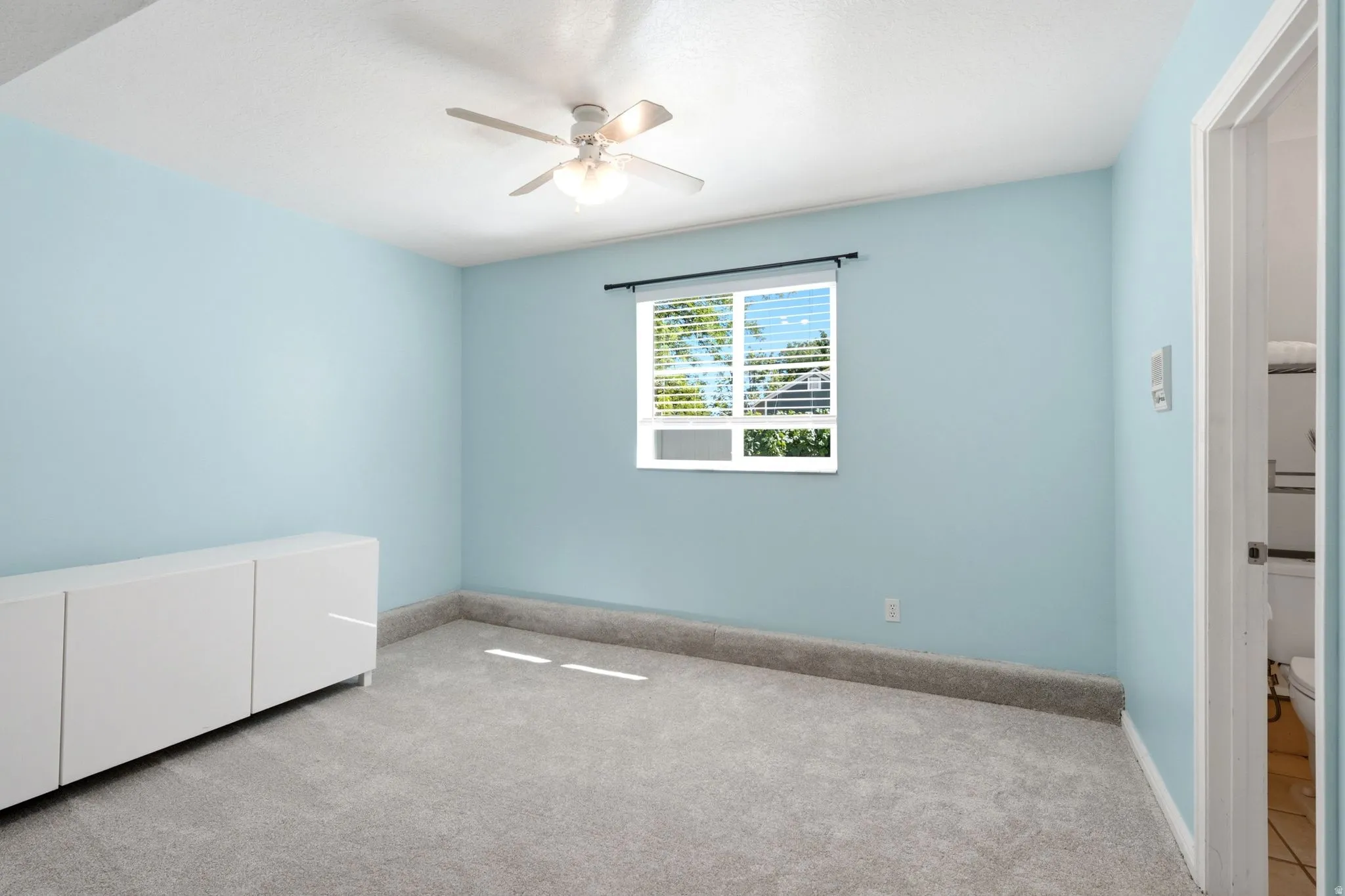 Bright room featuring a window with blinds, a ceiling fan, light blue walls, and grey carpeting