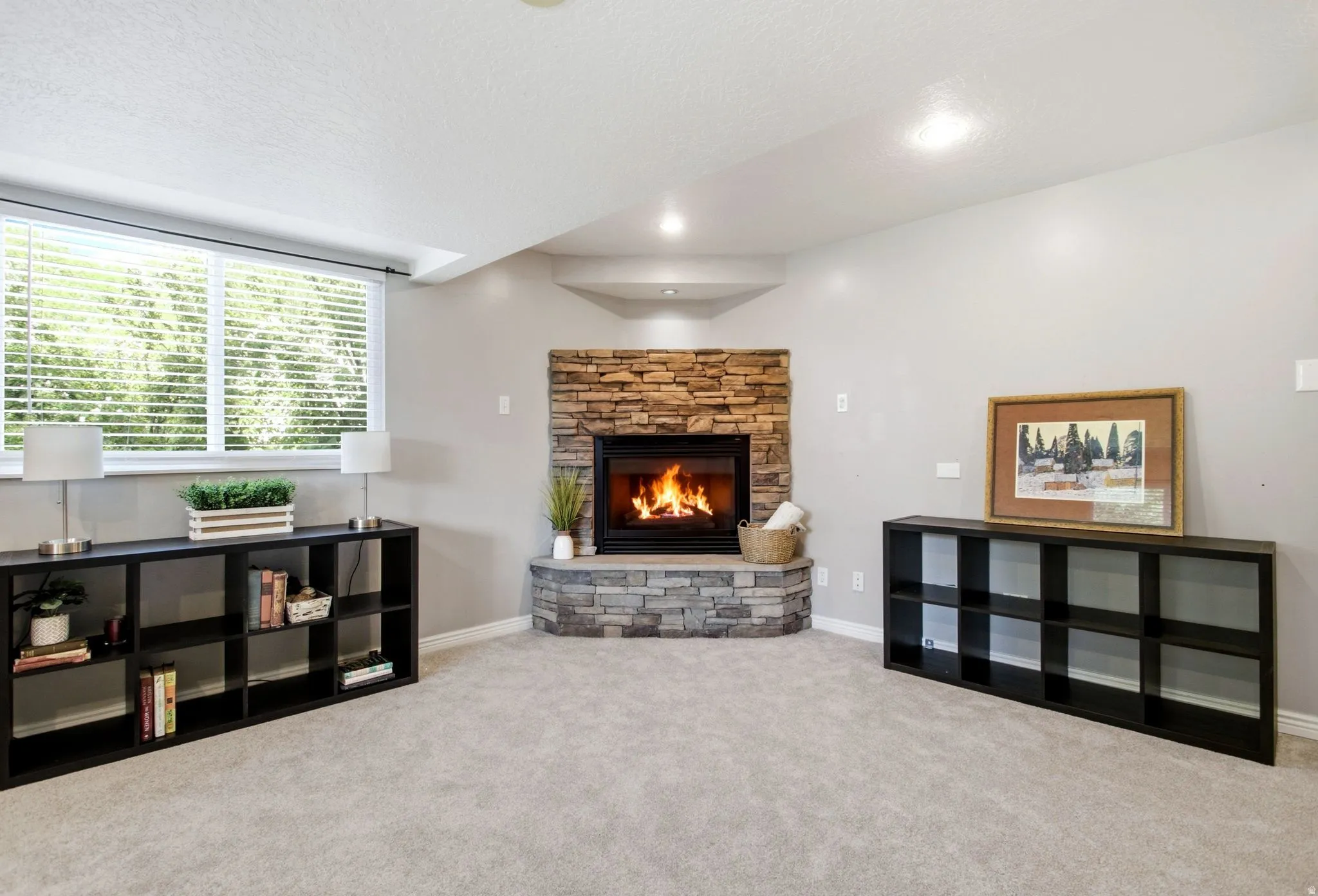Stone-clad corner fireplace, recessed lighting, expansive window with blinds, carpeted flooring, and light-toned walls