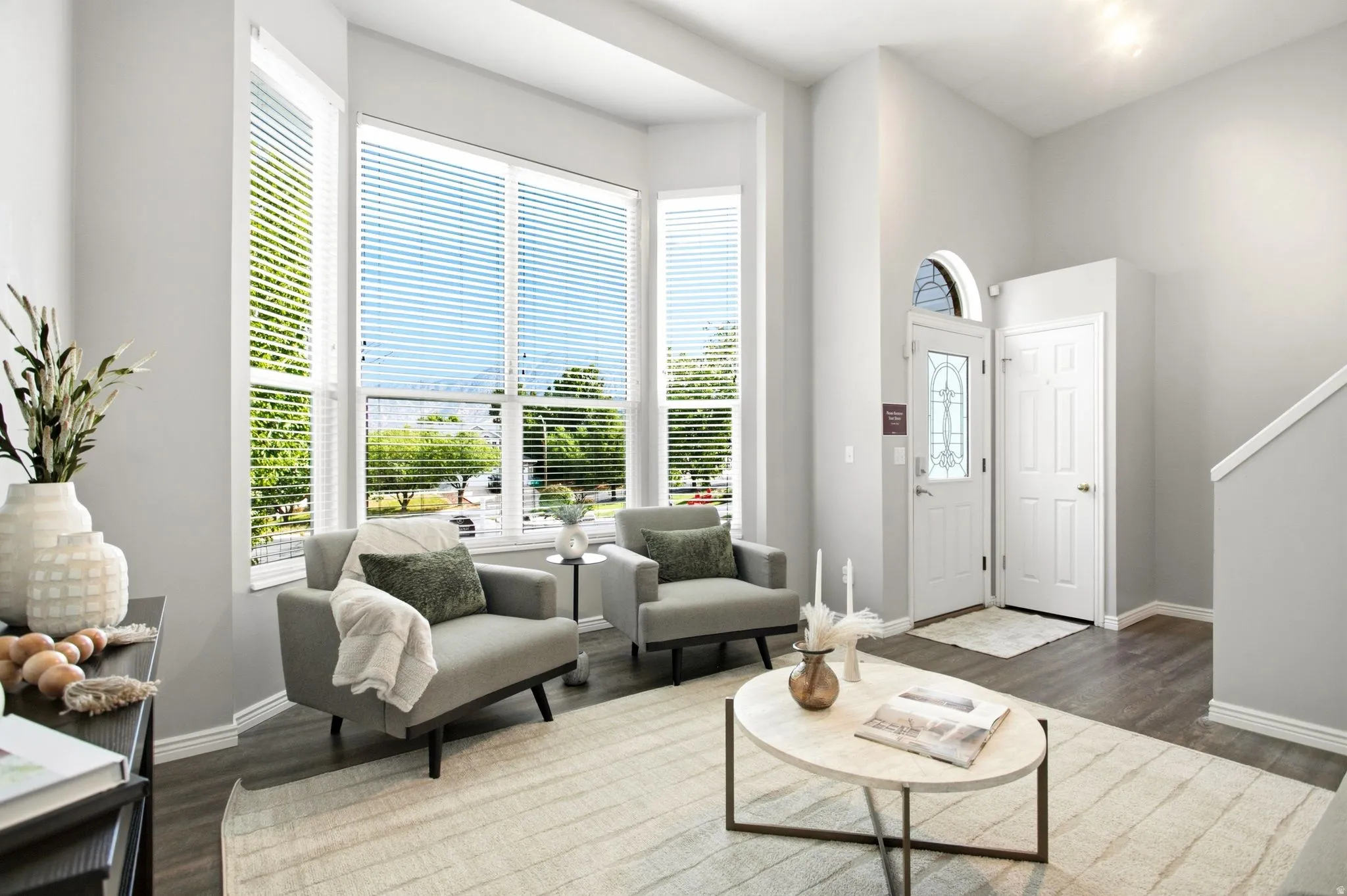 Bay window with horizontal blinds, wood-finish flooring, white entry door with decorative glass, arched transom window, and staircase with white risers