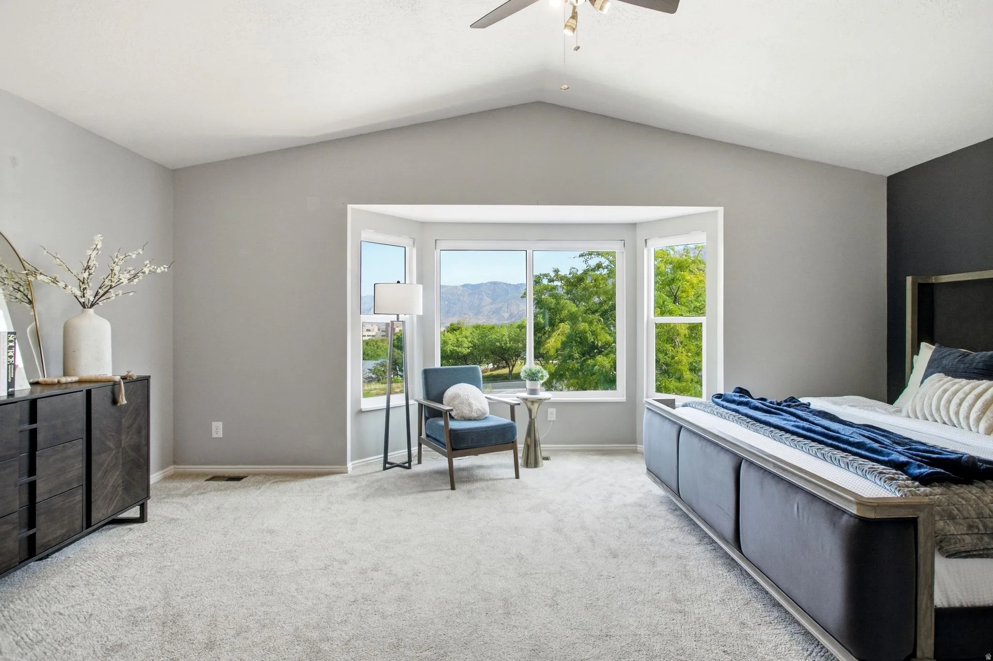 Vaulted ceiling with a ceiling fan, bay window, light grey carpeting, and dark accent wall