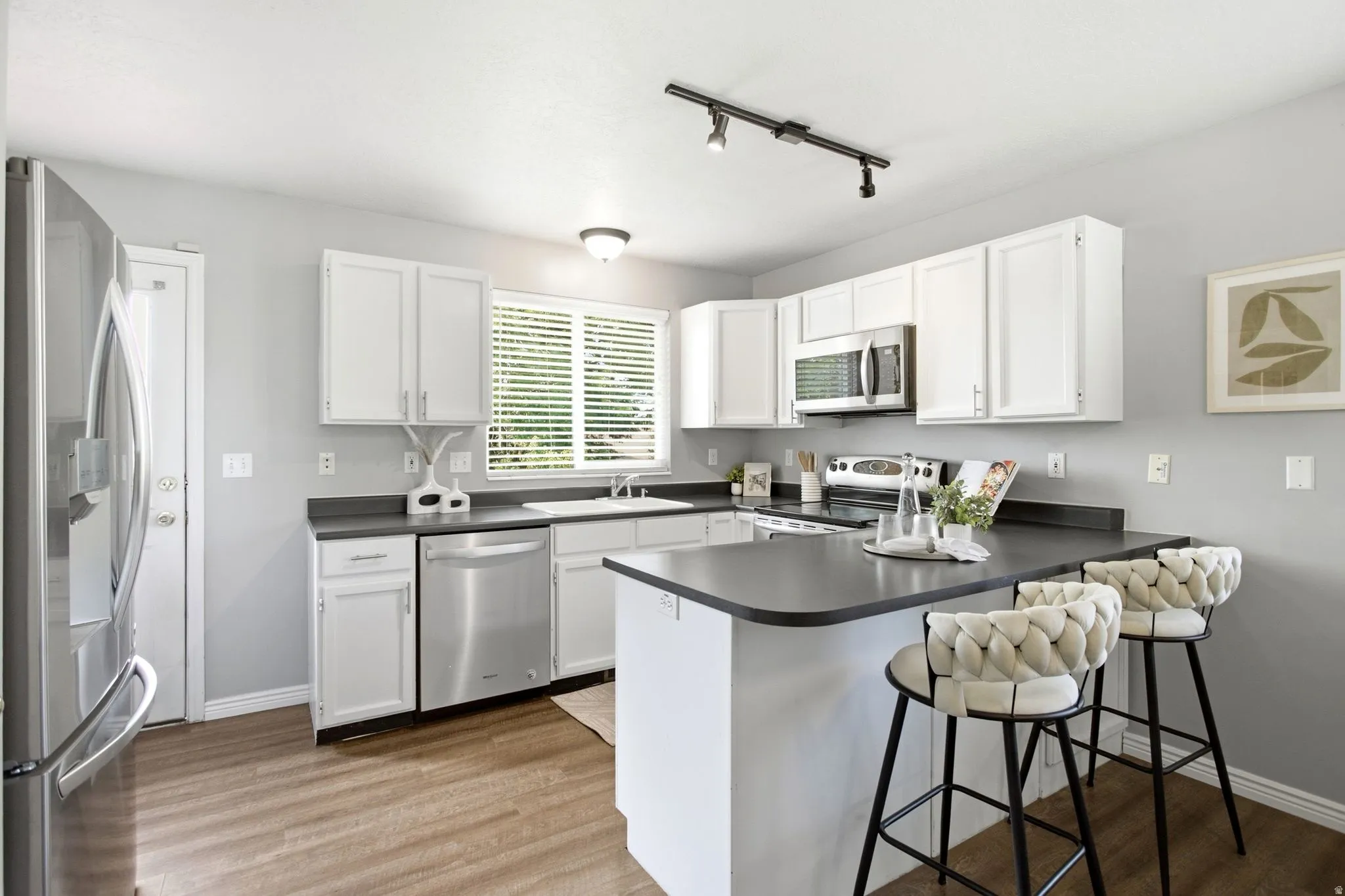 Modern kitchen featuring white cabinetry, stainless steel appliances, dark countertops, wood-finish flooring, and a breakfast bar