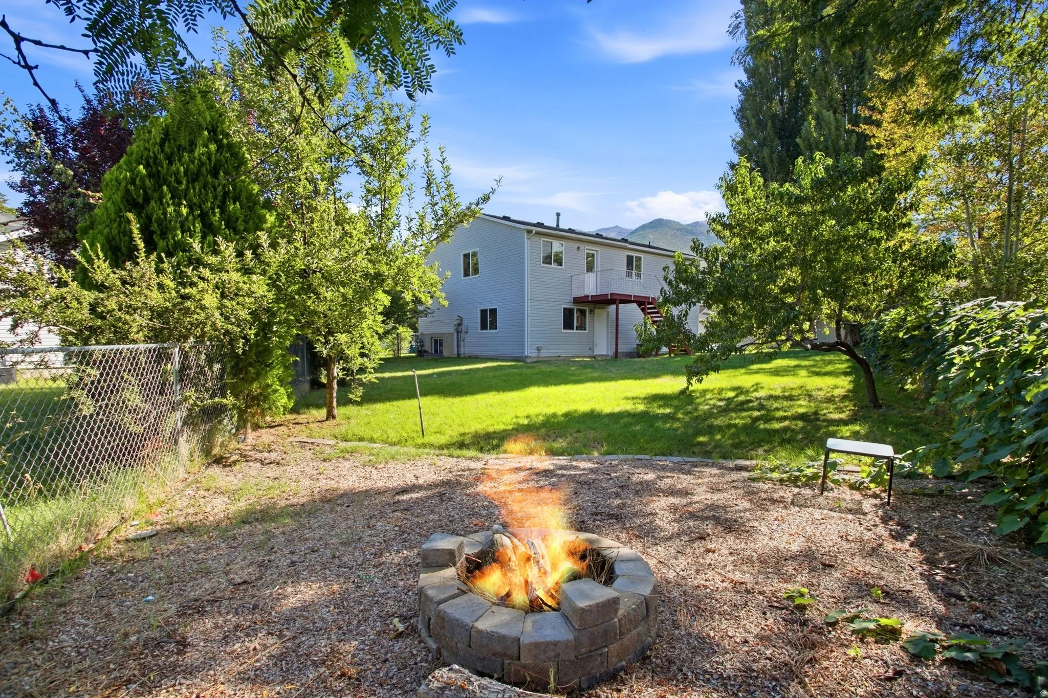 Expansive backyard featuring a stone-block fire pit, chain-link fencing, mature trees, and a multi-level structure with an exterior staircase