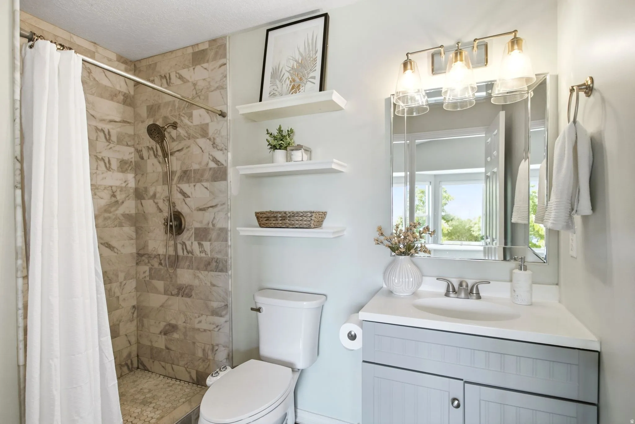 Shower with stone-finish wall tile, pebble-finish shower pan, wall-mounted shelving, corner vanity with undermount sink, and three-light vanity fixture