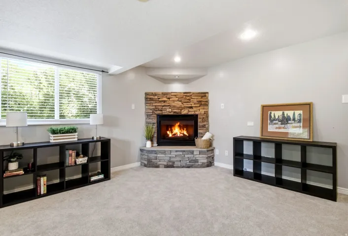 Stone-clad corner fireplace, recessed lighting, expansive window with blinds, carpeted flooring, and light-toned walls