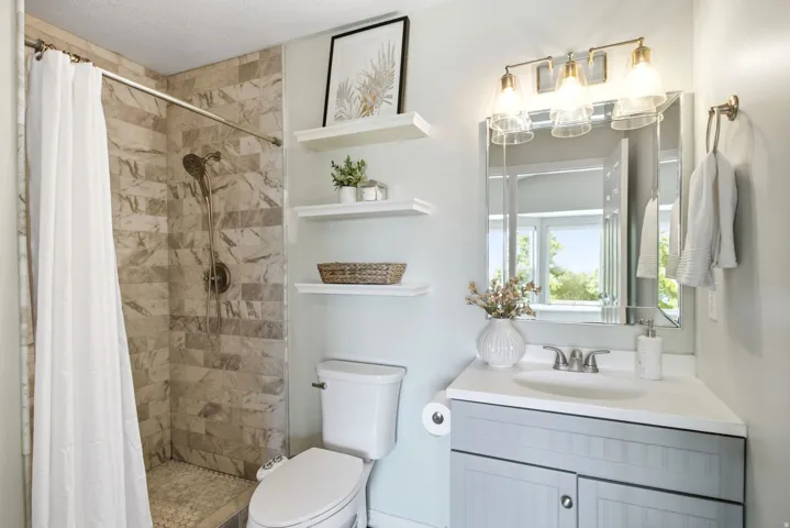 Shower with stone-finish wall tile, pebble-finish shower pan, wall-mounted shelving, corner vanity with undermount sink, and three-light vanity fixture