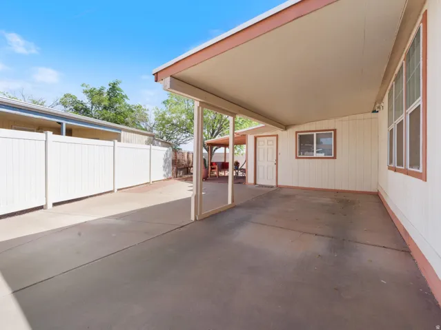 Expansive carport featuring a concrete surface, covered patio structure, multiple windows, a paneled door, and a white privacy fence