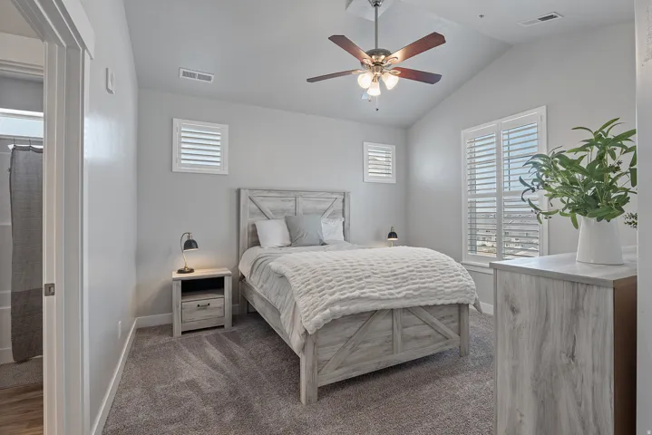 Bedroom featuring vaulted ceilings, ceiling fan with integrated lighting, textured carpeting, plantation shutters, and a rustic wood-finish dresser