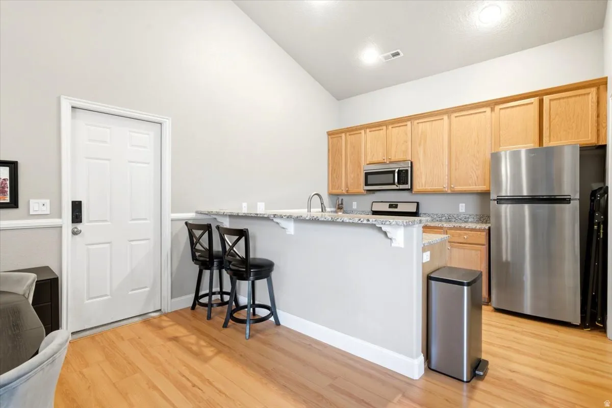 Kitchen featuring light wood cabinetry, stainless steel appliances, granite-style countertops, wood-finish flooring, and vaulted ceilings