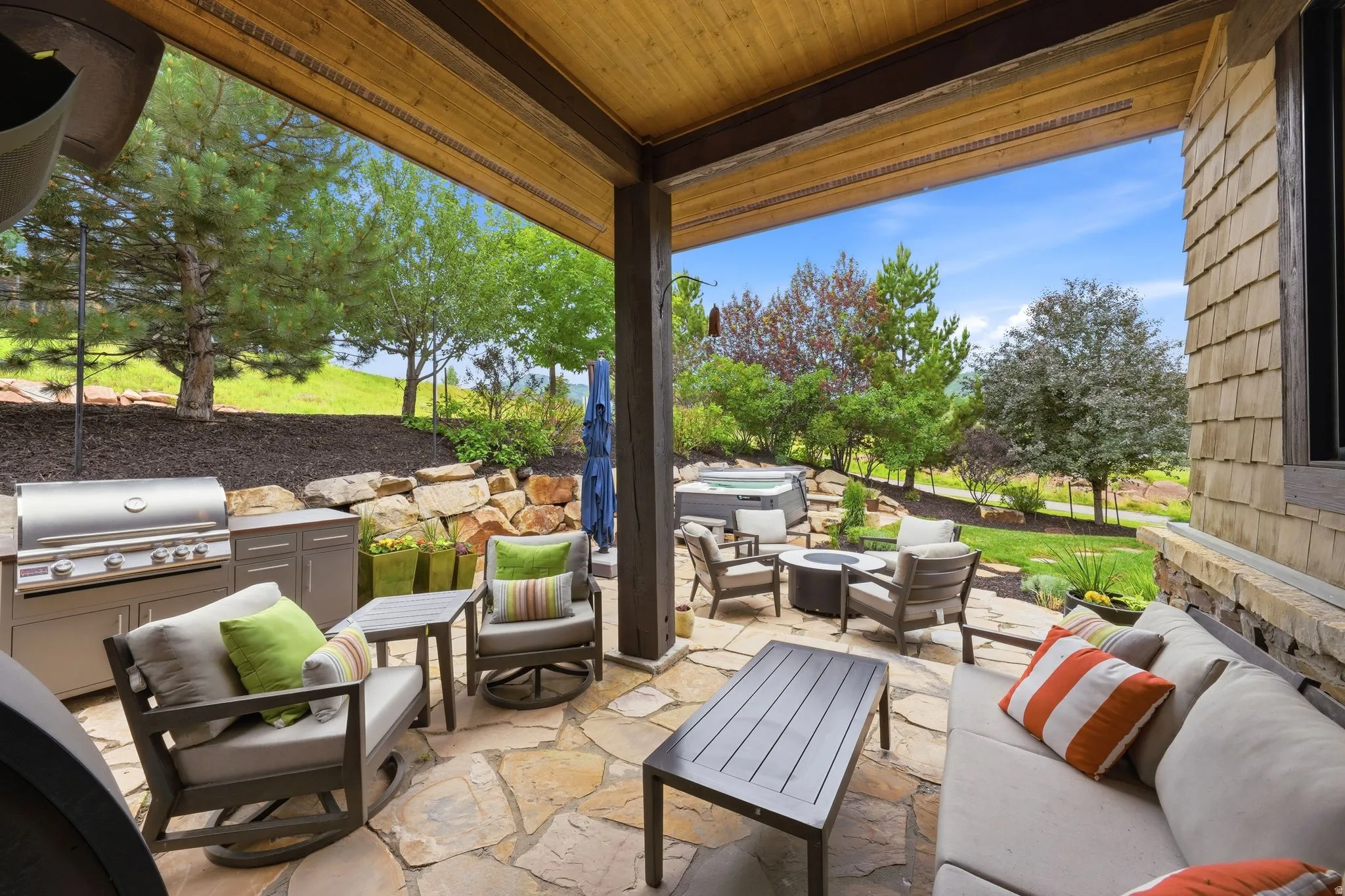 Covered outdoor patio featuring a flagstone surface, wood-paneled ceiling, built-in outdoor kitchen with grill, and stone retaining wall