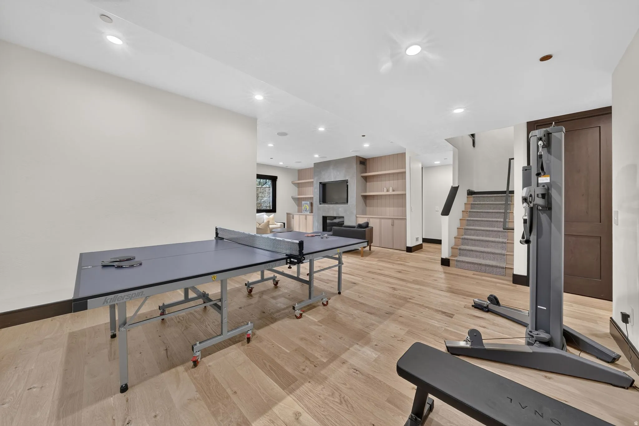 Expansive recreation room featuring light wood-finish flooring, recessed lighting, a built-in media center with shelving, and a carpeted staircase with a dark wood railing