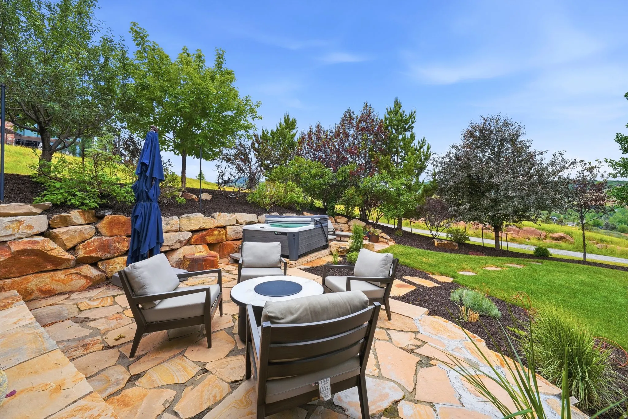 Flagstone patio featuring a hot tub, stone retaining wall, landscaped plantings, and a manicured lawn