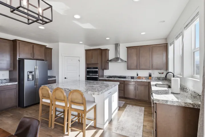 Spacious kitchen featuring a large central island with seating, extensive stone-finish countertops, stainless steel appliances, dark wood cabinetry, and multiple windows