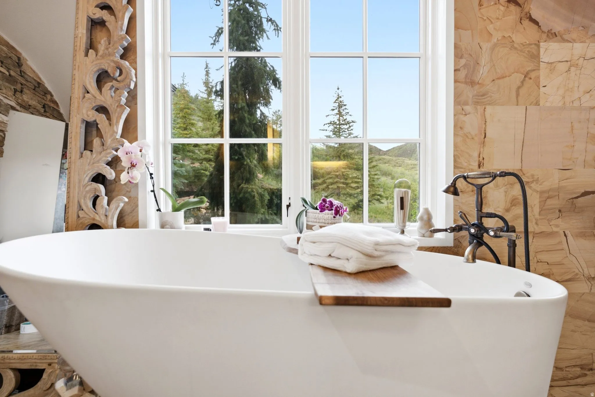 Freestanding soaking tub with an antique-finish tub filler, white window framing natural scenery, and a stone-tiled wall