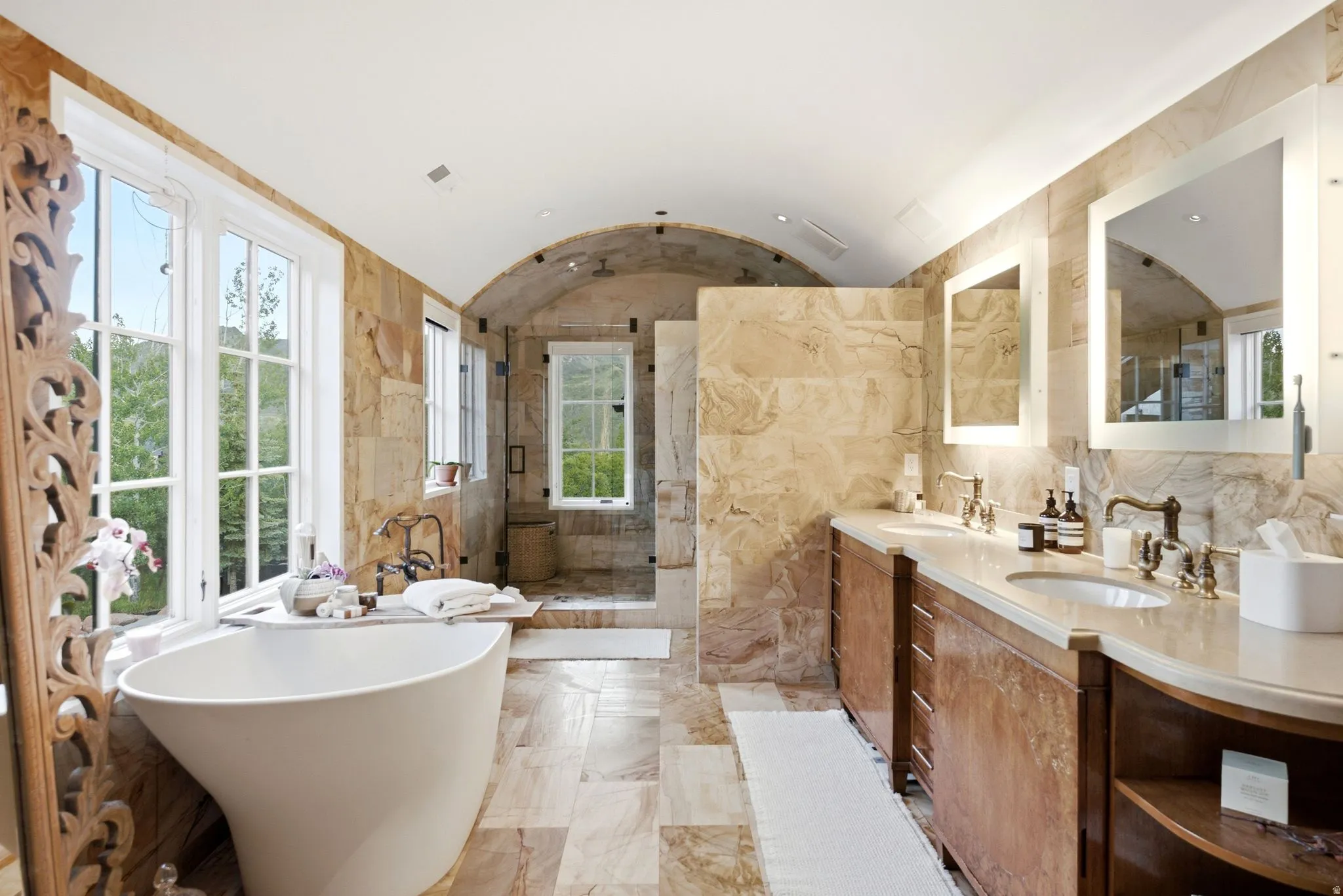 Spacious bathroom featuring a freestanding soaking tub, dual vanity with undermount sinks, stone tile flooring, arched ceiling, and large windows