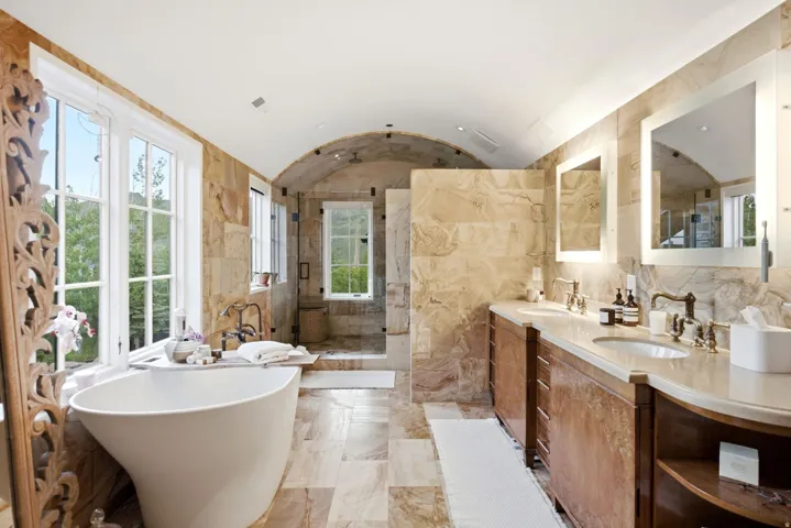 Spacious bathroom featuring a freestanding soaking tub, dual vanity with undermount sinks, stone tile flooring, arched ceiling, and large windows