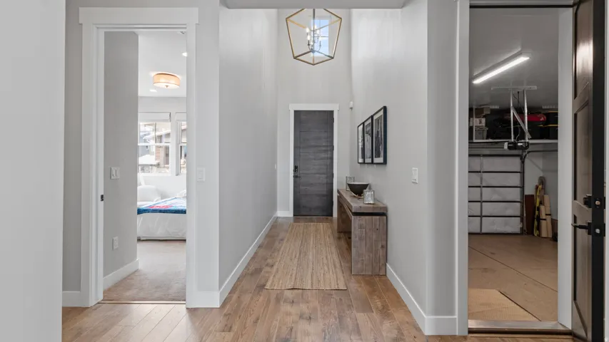 Entry hallway looking towards the front door.  Wood-finish flooring, a contemporary geometric chandelier, and tall ceilings.  The second bedroom and second bathroom are located to the left while the garage and laundry are to the right.