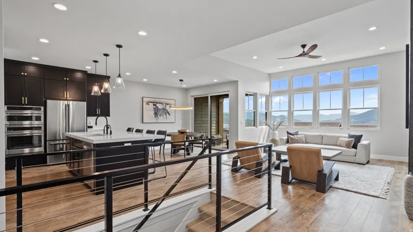 Open-concept living space with wood-finish flooring, a contemporary kitchen featuring dark cabinetry and stainless steel appliances, a center island with seating, a dining area, and a living room with a prominent wall of windows