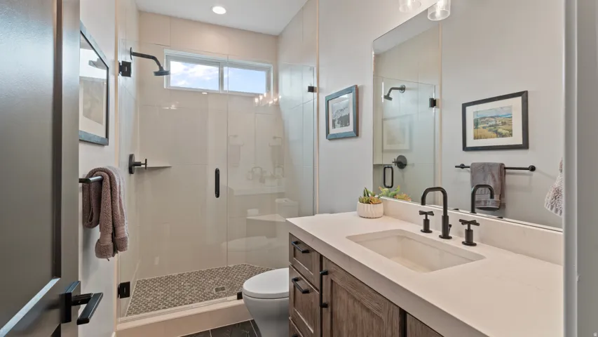 Main level bathroom (Bathroom #2) featuring a spacious glass-enclosed shower with mosaic tile flooring, a custom wood-finish vanity with an integrated sink, matte black fixtures, and a transom window for natural light