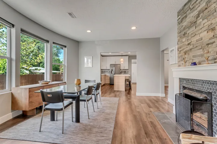Spacious dining area featuring wood-finish flooring, a stacked stone fireplace with a hexagon tile surround and white mantel, and large windows providing natural light