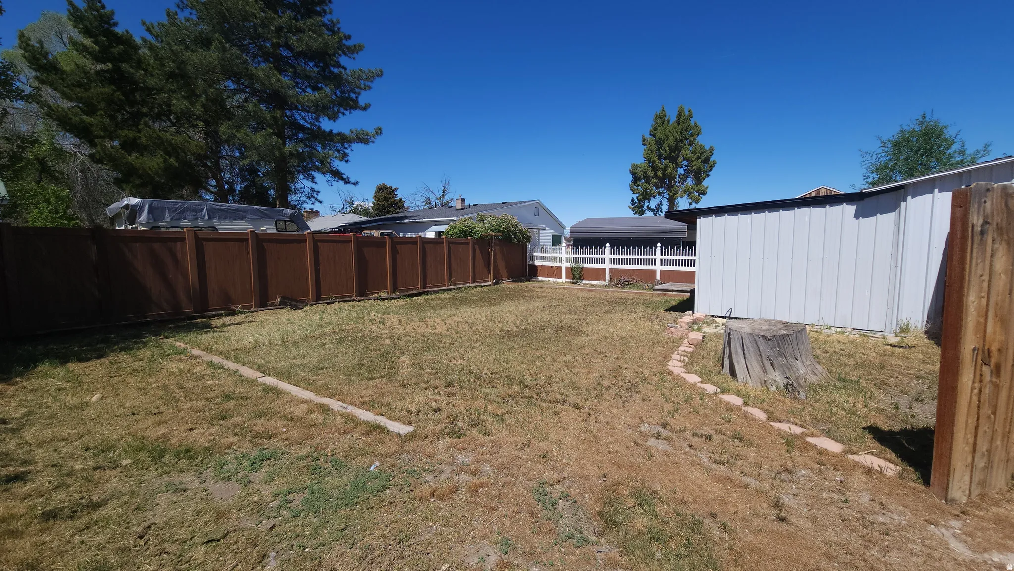Expansive backyard area featuring a brown privacy fence, a white metal outbuilding, and a decorative stone border