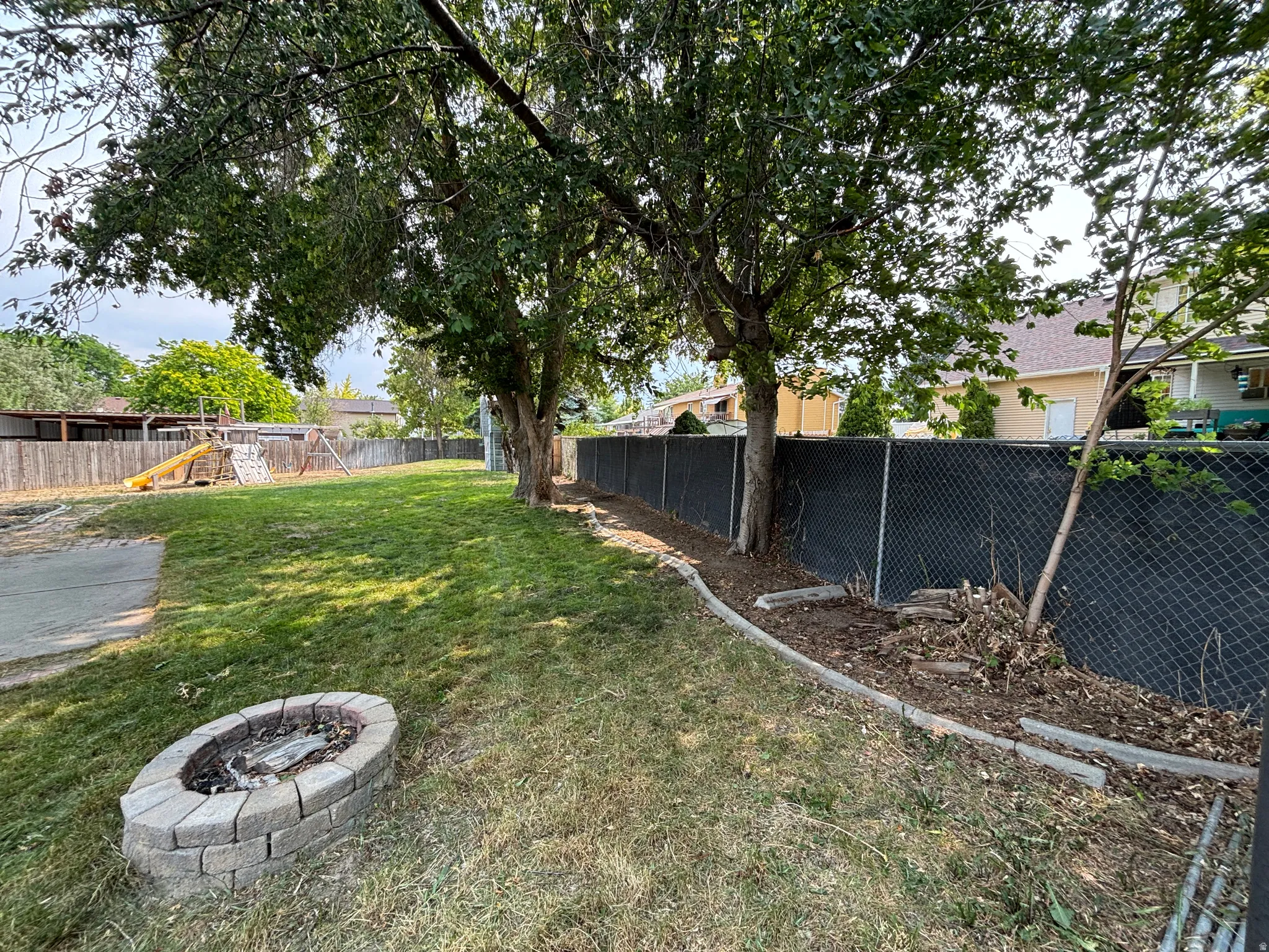 Expansive backyard featuring a stone-lined fire pit, mature trees, and a chain-link fence