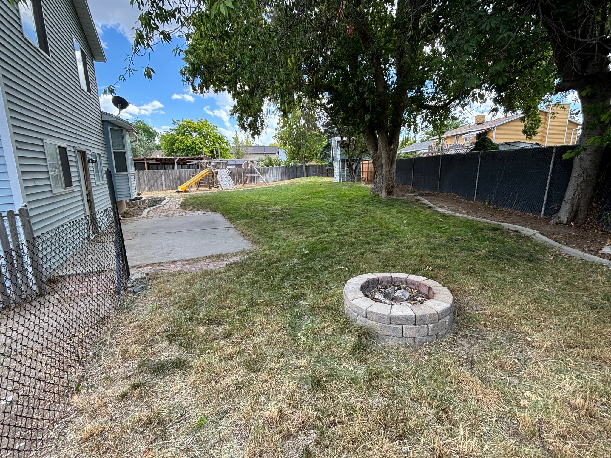 Expansive backyard featuring a stone block fire pit, mature trees, and a concrete patio pad