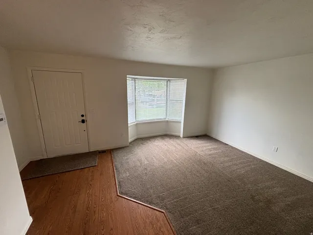 Living area with a prominent bay window, neutral wall finishes, and a combination of wood-finish flooring and carpeting
